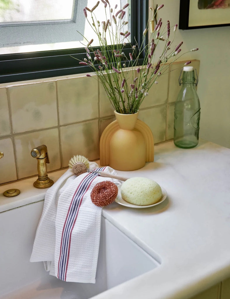 The Tradition Dish Kit by Andree Jardin includes a woven white dish towel with blue and red stripe, a metal scrubber, and a wooden scrubber, and sits next to a bar of soap and a modern vase holding cut stems next to a sink.