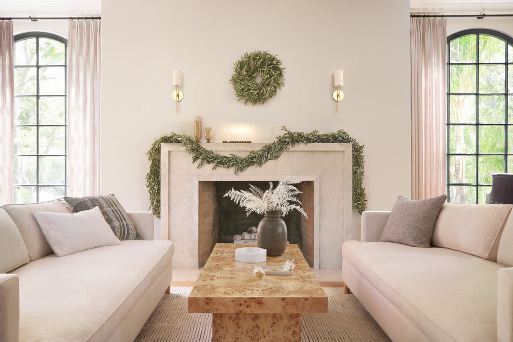 A light stone fireplace is draped in rustic garland. In front of the fireplace are two modern cream sofas and a burl wood coffee table.