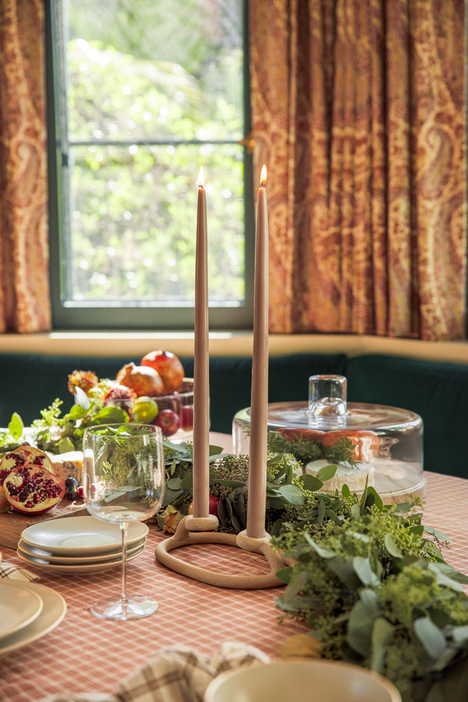 The SIN Duo Candlestick holder holds two lit tapered candles atop a holiday dining table, surrounded by green garland and a glass cloche on a red checked tablecloth.