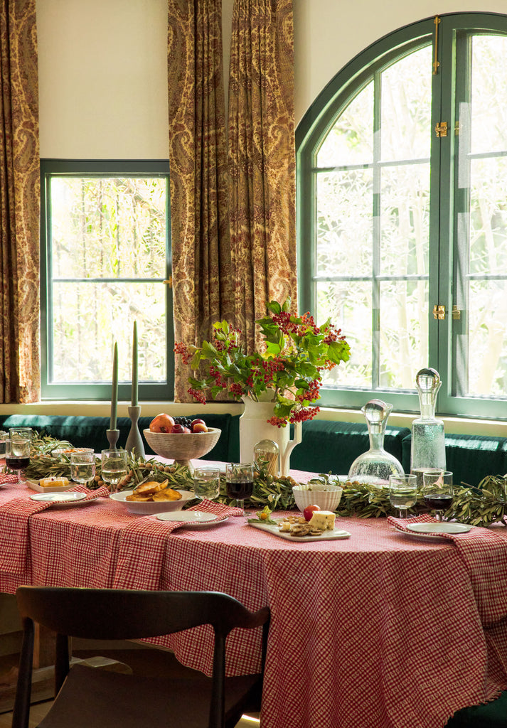 A holiday table is set with a red plaid tablecloth over an oval table set into a banquet. Fruits, cheeses, and snacks are set out along with glasses of wine and a large white milk jug filled with red berry sprigs.