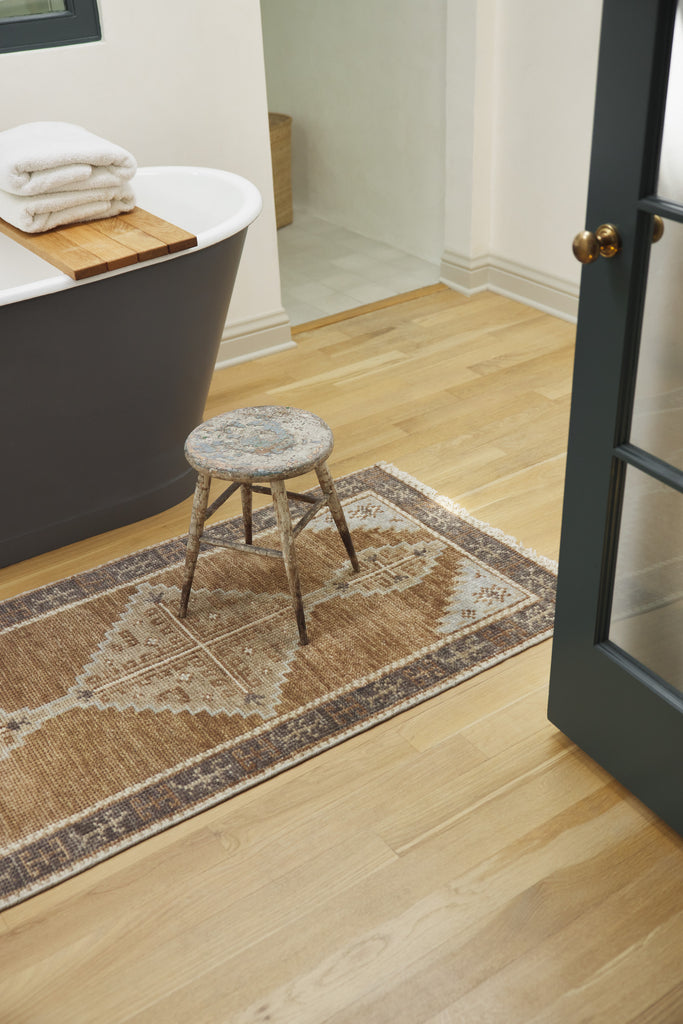 A traditional neutral and brown earth toned small rug sit in front of free-standing black bathtub. A small weathered foot stool sits on the rug.