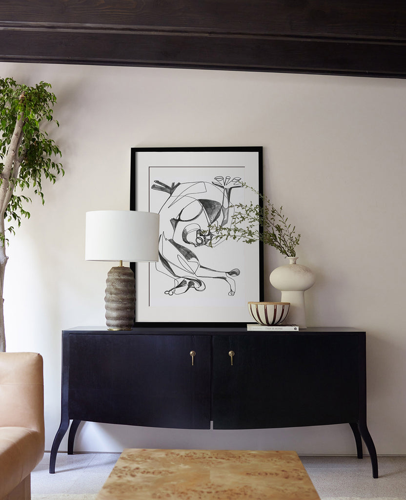 A black and white sketch by artist Adrian Brandon leans on top of a black sideboard cabinet. A lamp and two vases flank the print.
