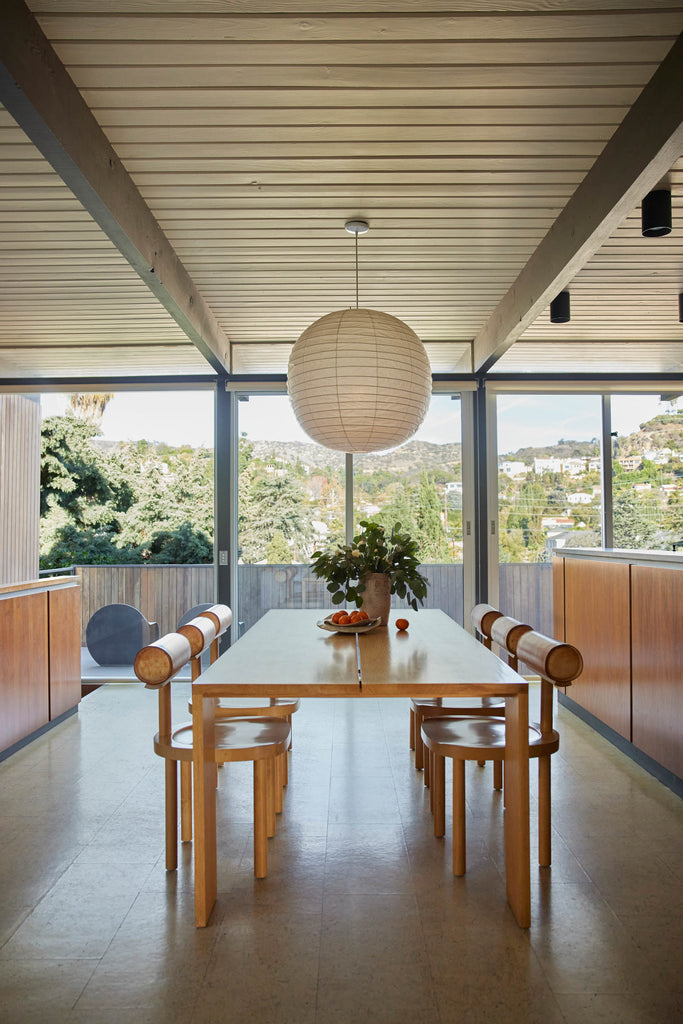 Emilie Halpern's modern dining room includes a round globe paper lantern hanging above a long wooden rectangular dining table with six modern wooden open-backed dining chairs with round backs sitting across from each other.