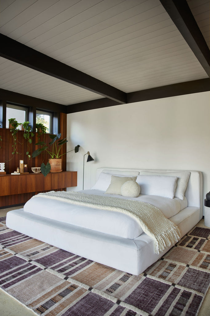 Emile Halpern's mid-century modern bedroom includes a white thick padded upholstered bed with white and cream bed linens, throw pillows and throw blanket, atop a brown, tan and neutral grid patterned area rug.