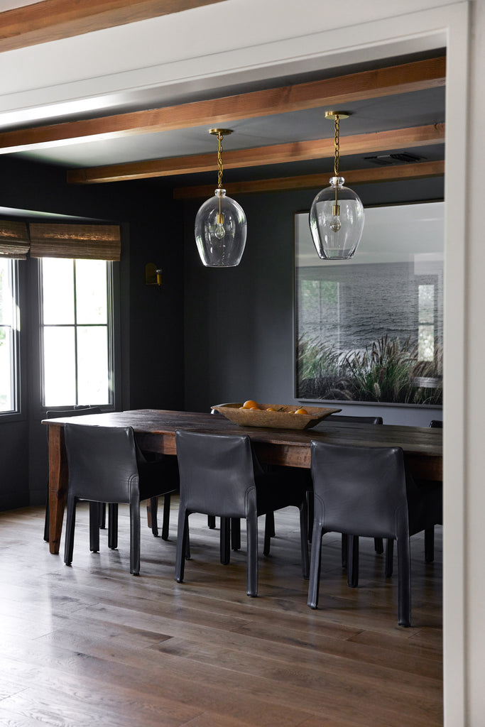 The dining room of Sarah Solis' home is painted black and has black leather dining chairs surrounding a long dark wood table. A long square bowl of fruit sits on the table and two glass pendant lights hang above it.
