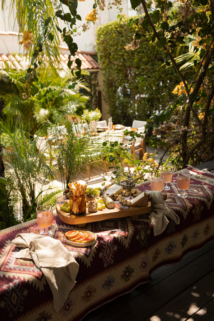 A charcuterie board and four cocktail glasses sit on a southwestern fabric-wrapped bench overlooking an outdoor dining area.