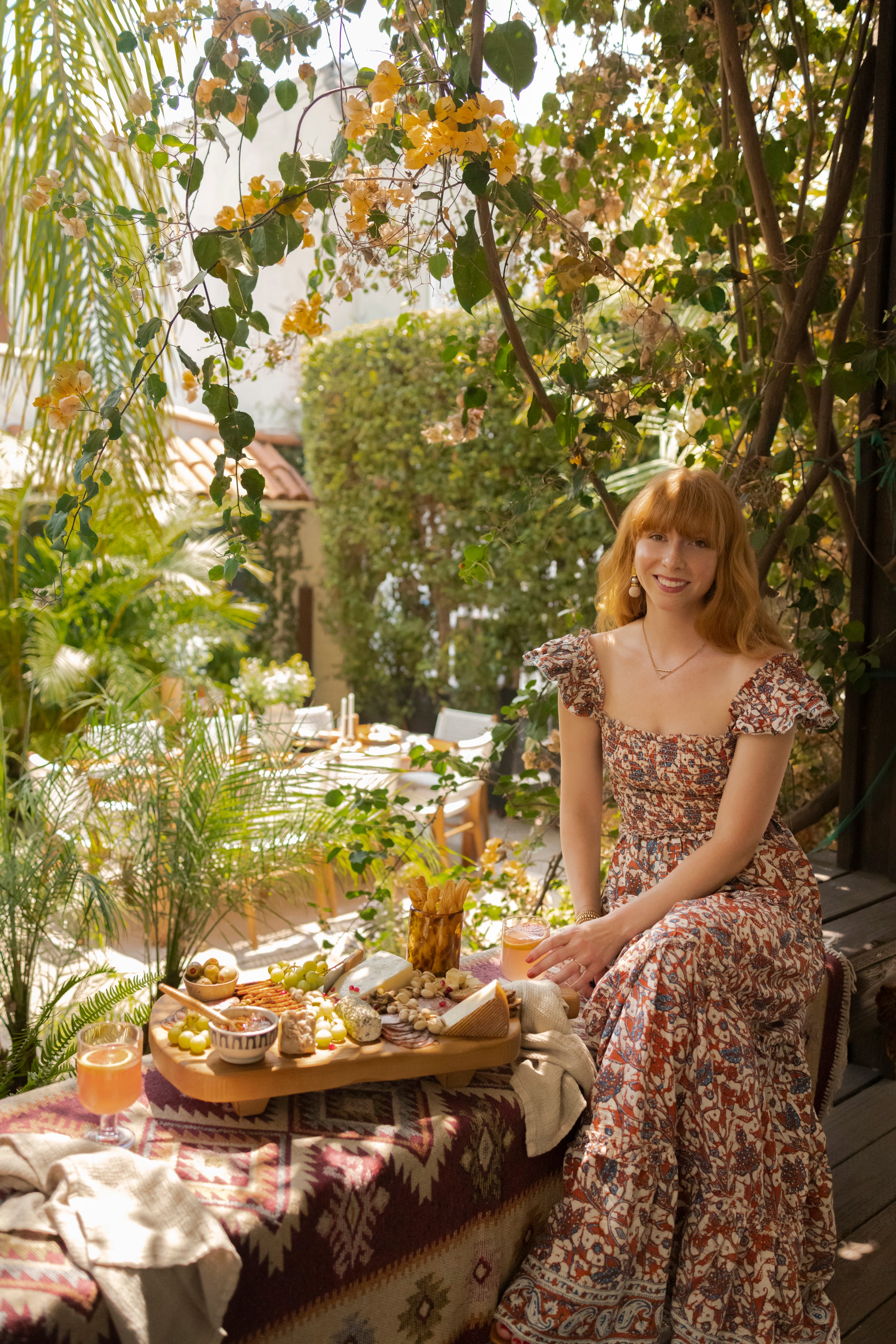 Interior designer Lexie Layne Sokolow wears a floral dress and sits on a bench next to a charcuterie board and cocktail glass overlooking an outdoor dining area.