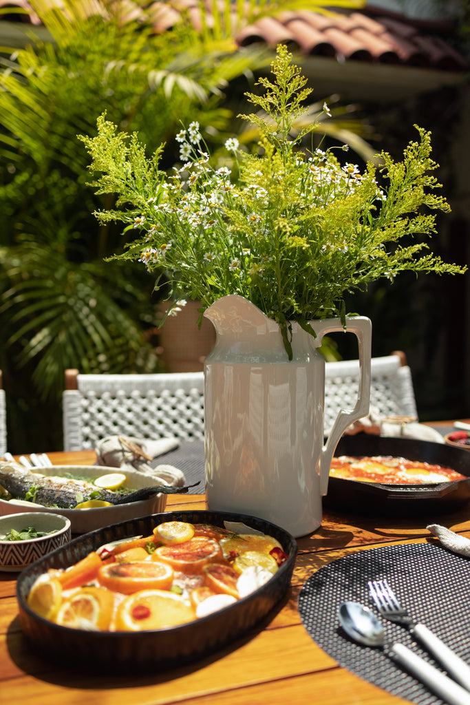 A large white pitcher full of greens and florals sits on an outdoor teak dining table surrounded by serving bowls and platters of salad and fish.