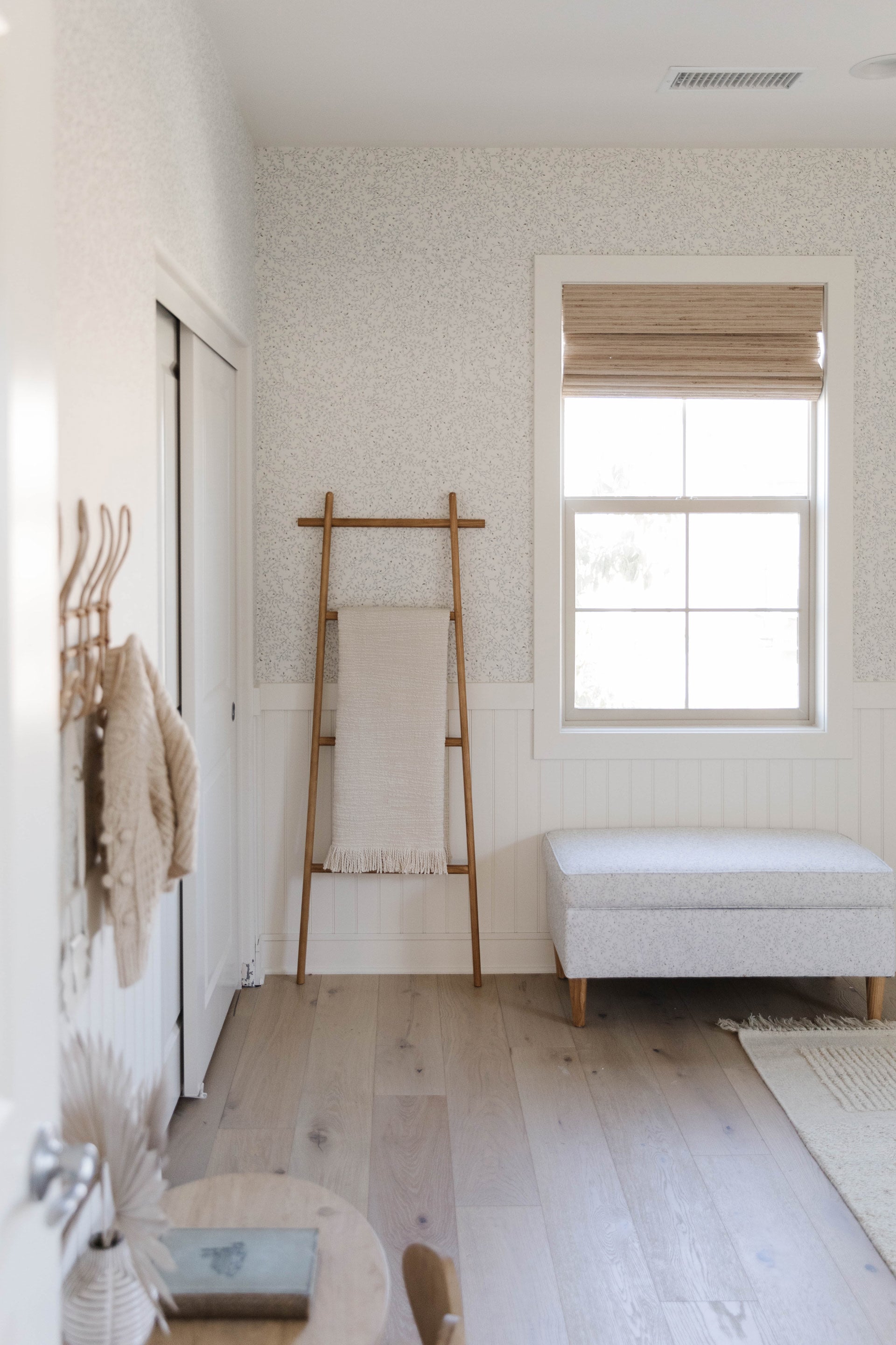 The Dainty Leaves botanical wallpaper sets the tone for this room with a light wood blanket ladder and a small light gray upholstered ottoman.