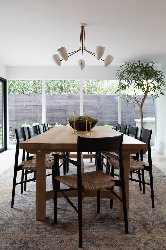 A modern dining room has a long wooden dining table surrounded by ten black wood chairs with woven seats and a modern chandelier above.