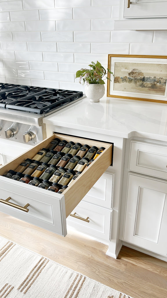 A white kitchen cabinet drawer is open, revealing rows of labeled cooking herbs and spices laying flat in glass jars.