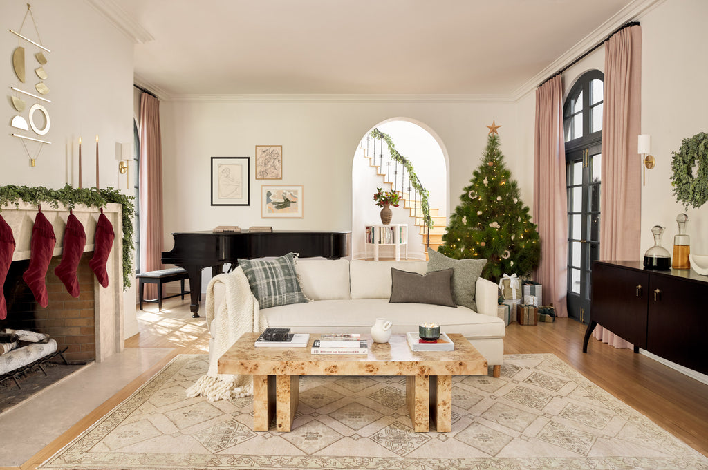 The gold minimalist Quadrant wall hanging from Circle & Line hangs above a holiday fireplace with garland, lit candles and deep red velvet stockings. The living room has an ivory sofa with neutral throw pillows and a Christmas tree surrounded by presents is in the corner.