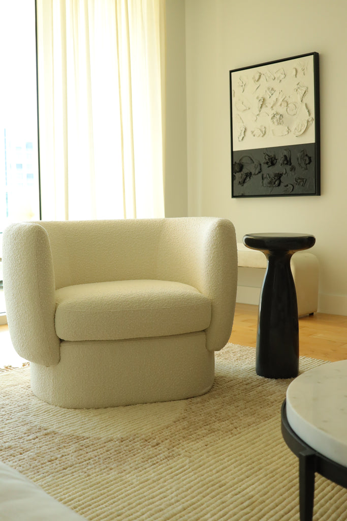 A light boucle rounded Zaha arm chair sits next to a small round black Marielle side table. A minimalist black and white piece of art hangs on the wall behind the chair.