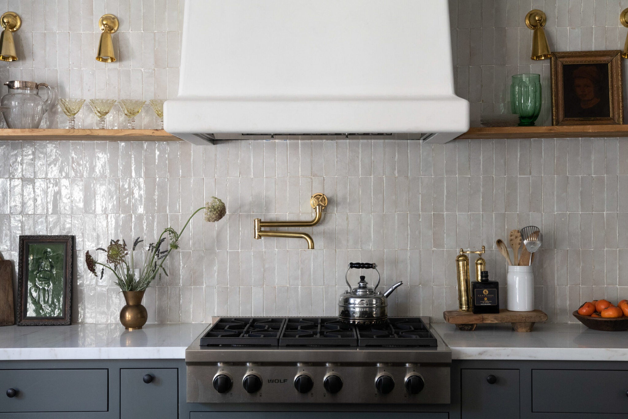 A stainless steel range  with a silver tea kettle on it has a pearlized stacked vertical tile backsplash behind it and a white vent hood and wooden open shelving on either side.