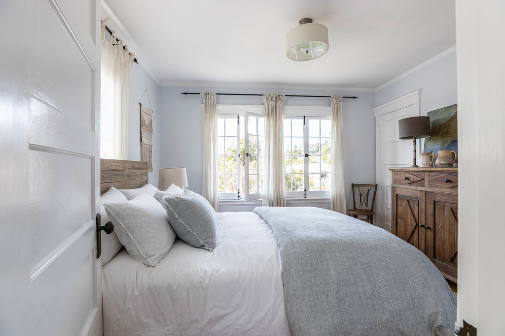 White bedding with a gray duvet and accent pillow sits on a bed with a light wood headboard. Across from the bed is a tall wooden dresser with a lamp, two tan pitchers and a leaning piece of art. A small wooden chair sits in the corner.