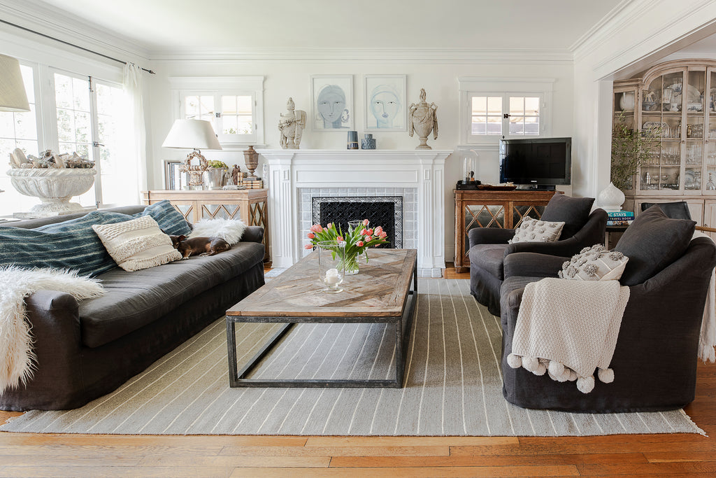 A dark sofa with neutral pillows, a small sleeping brown dog, a blue throw blanket and a fuzzy throw sit across from two dark upholstered armchairs with Pom Pom at home neutral throw pillows and a blanket. A wood and metal coffee table sits in the middle and a white fireplace is behind the table.