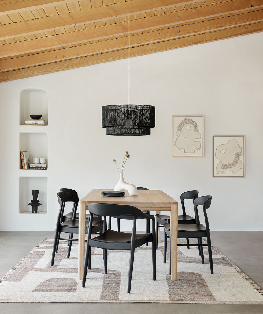 An Elan Byrd-designed neutral rug lays below a light wood dining table surrounded by six black curved wooden chairs. Two abstract prints hang on the wall behind the table.
