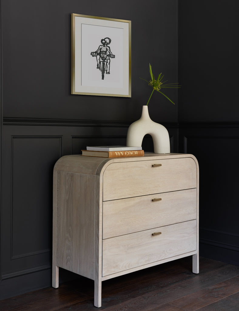 A gold framed Ride print by Adrian Brandon hangs above a curved light wood dresser offset by black walls.