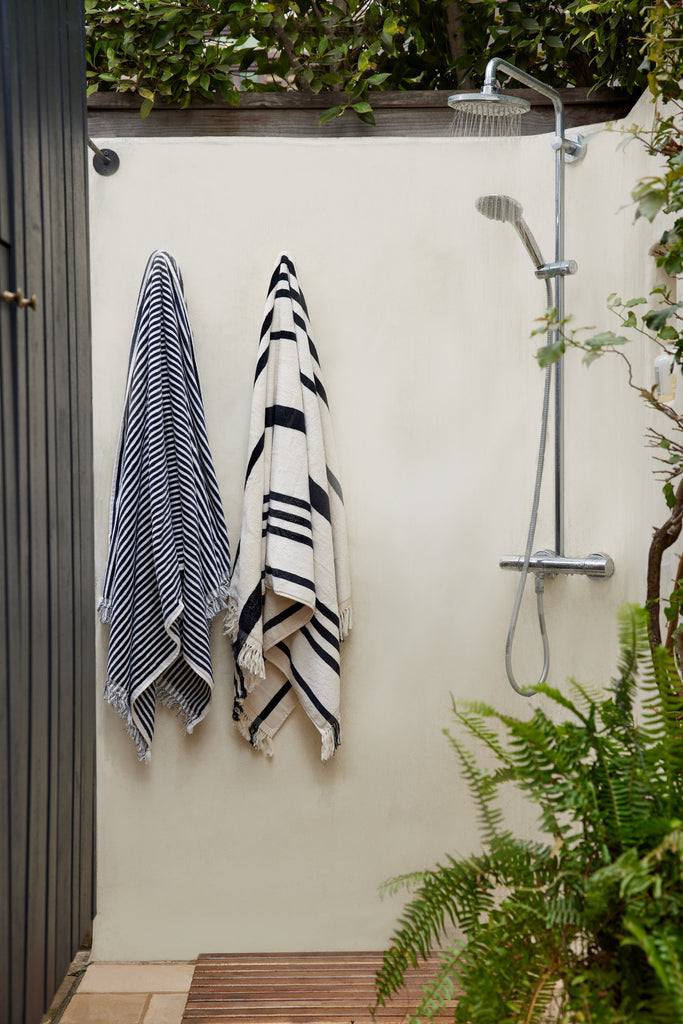 A navy and white striped and black and white striped towel hang in an outdoor shower area surrounded by ferns and plants.