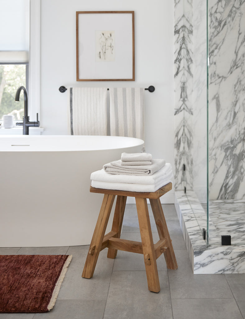 A small teak wood stool with a stack of towels on it stands in between a marble shower and a free-standing bathtub.