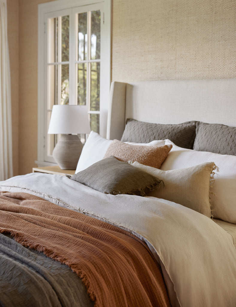 Neutral rust, brown, and cream bedding is layered on a bed with a neutral linen tall headboard.