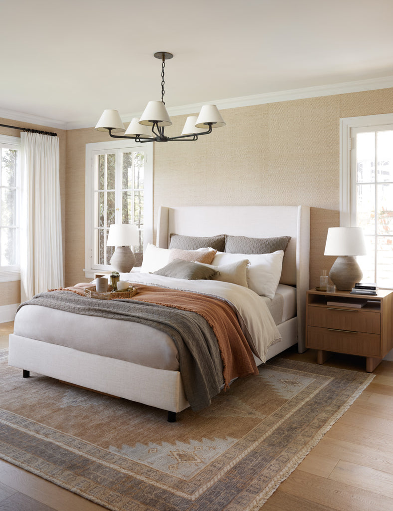 A neutral linen upholstered bed is layered with rust, brown, and cream bedding atop a neutral patterned area rug.