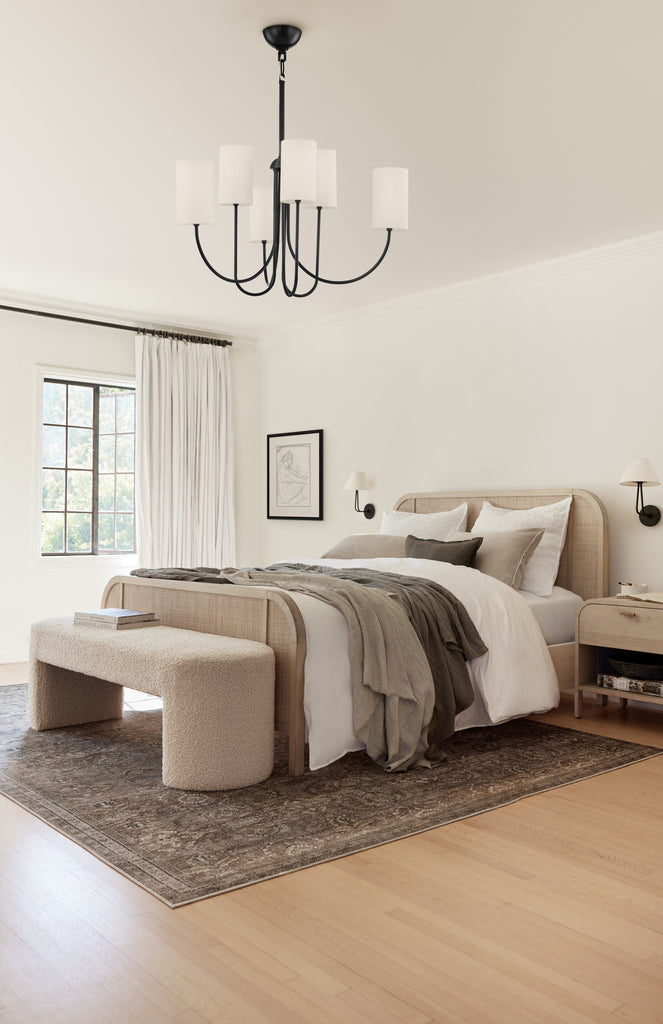 A light wood and cane bed with neutral bedding and dark gray blankets sit in this neutral bedroom. A rounded boucle bench sits at the end of the bed, which sits upon a dark traditional area rug. A slim black chandelier hangs above the bed.