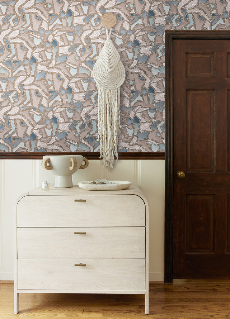 The blue and neutral Mosaic Wallpaper designed by Malene Barnett has a metallic and geometric design. A crocheted wall hanging hangs on the wall above a light wood curved dresser.