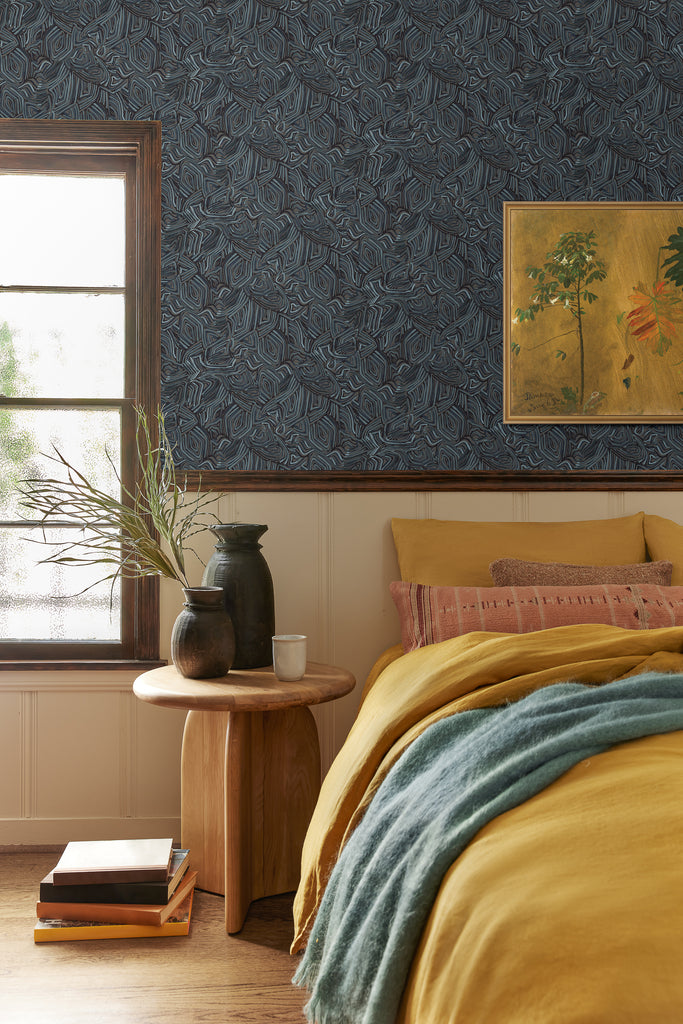 The dark blue Bequia Wallpaper designed by Malene Barnett hangs in a colorful bedroom and contrasts against off-white wainscotting. Against the wall is a bed covered in golden bedding, a blue throw blanket and rose throw pillows.