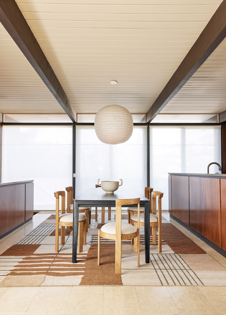 A dark wood rectangular dining table is surrounded by six light wood curved modern dining chairs with ivory seats. A round paper globe pendant light hangs above the table, which sits atop a neutral and brown geometric grid area rug.