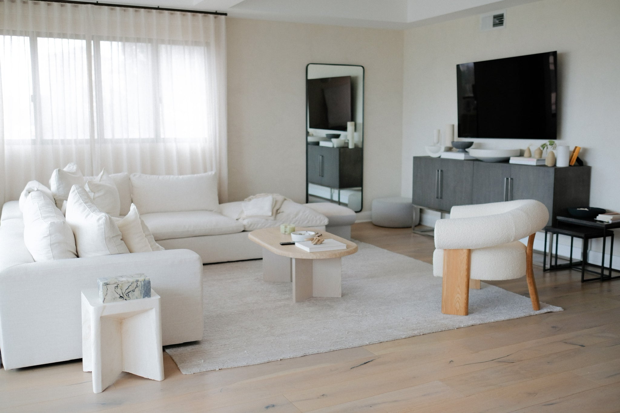 A low white sectional sofa with loose back cushions sits in a white living room with a short white square end table, oval light wood coffee table, a curved white boucle accent chair and a black media cabinet.