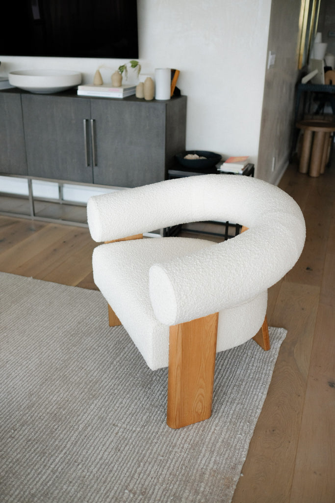 A curved white boucle arm chair with wood legs sits on a neutral striped area rug.