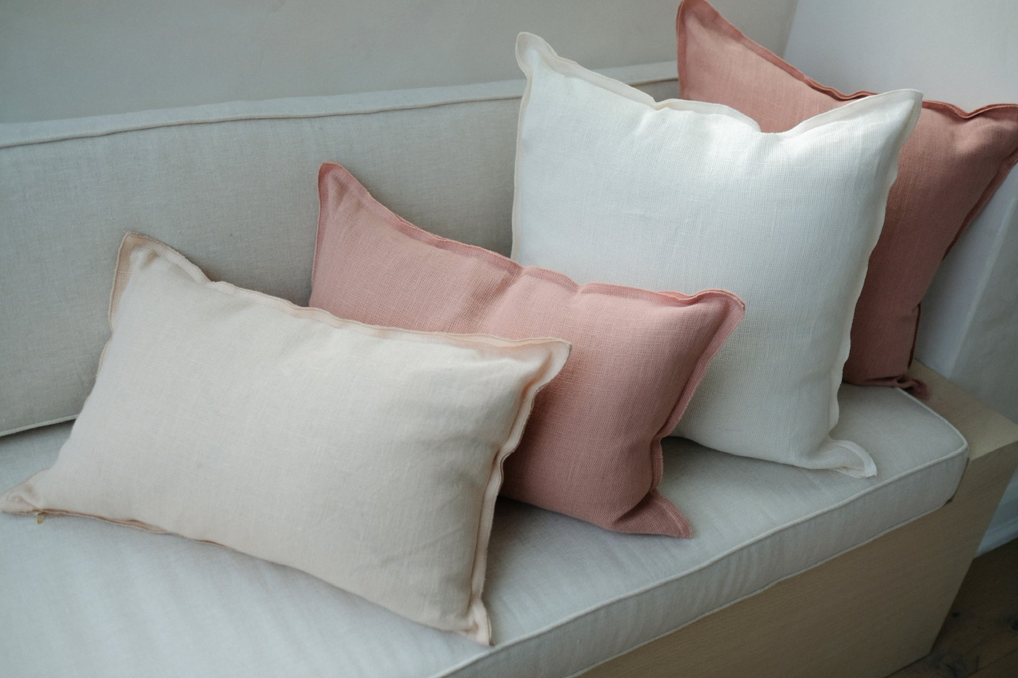 A cream linen and a rose linen lumbar pillow layer with one ivory linen and one rose linen square throw pillows on a neutral colored linen upholstered bench.