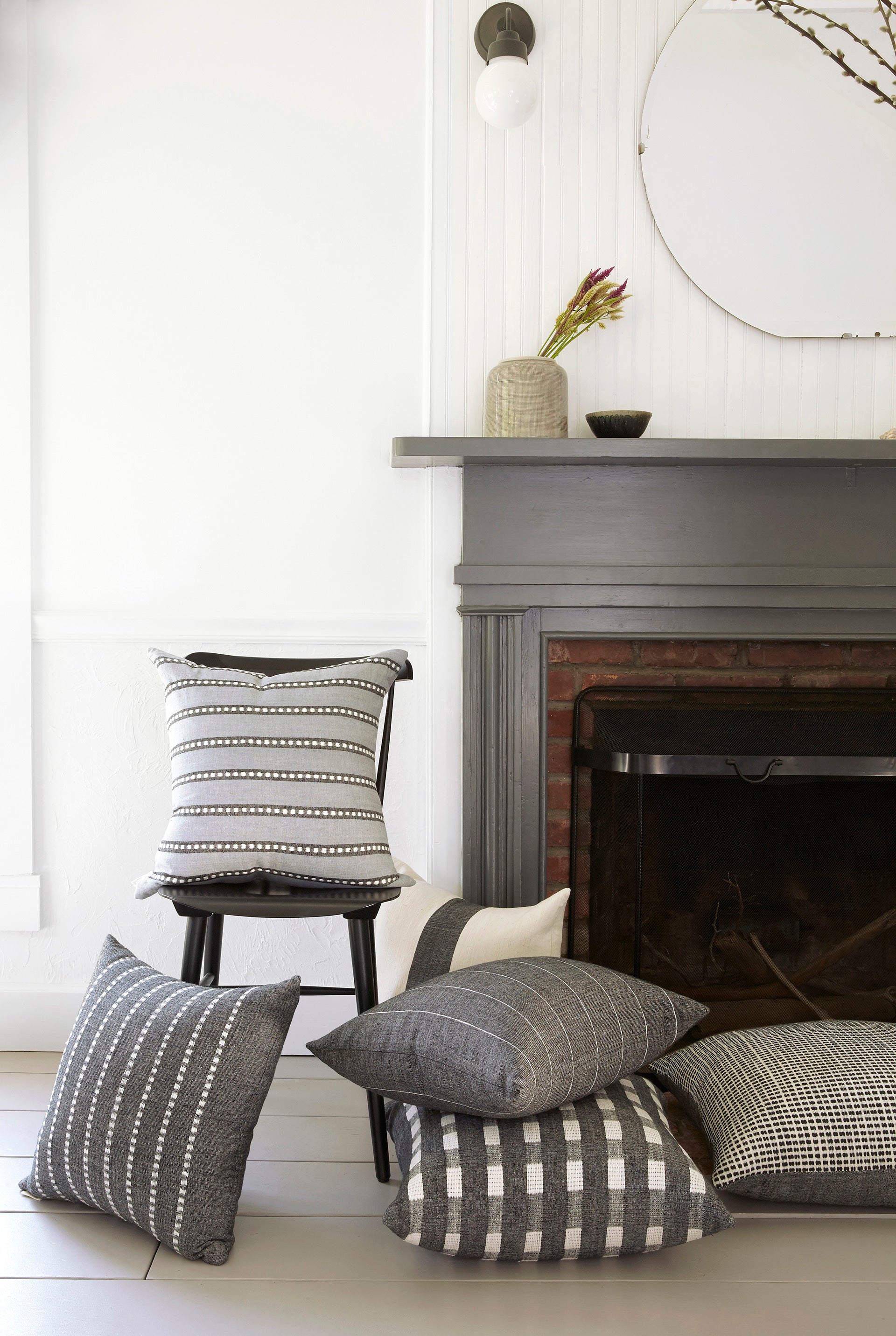 A stack of gray, black and white Bole Road throw pillows are piled on  the floor and a small wooden chair next to a gray fireplace.