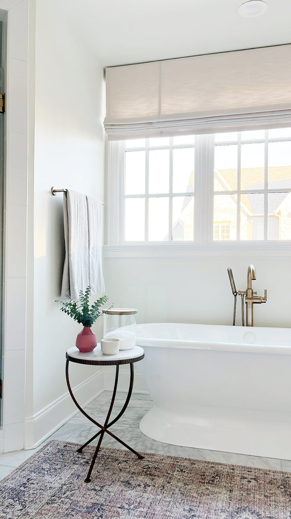 A free standing bathtub sits below a window and has a free-standing gold faucet. A small round table with black legs and a white top hold bath salts. Two white towels hang to the side.