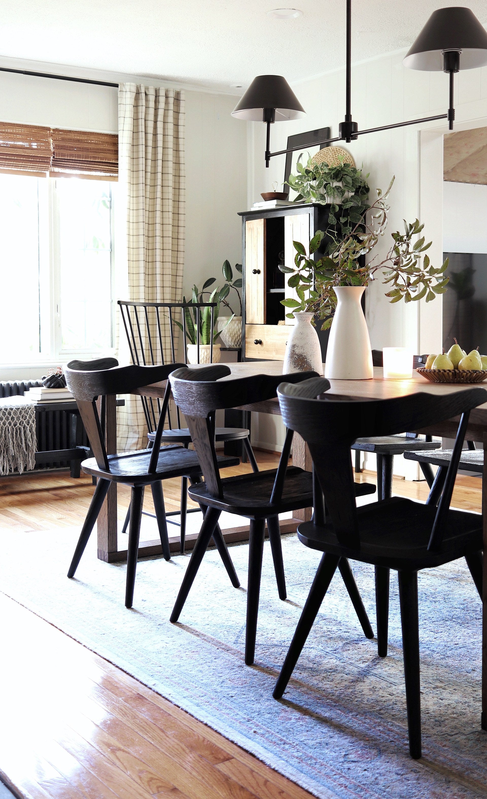 Black wooden dining chairs surround a wooden rectangular dining table. Two tall white rustic vases hold tree clippings and a basket of pears sits next to them.