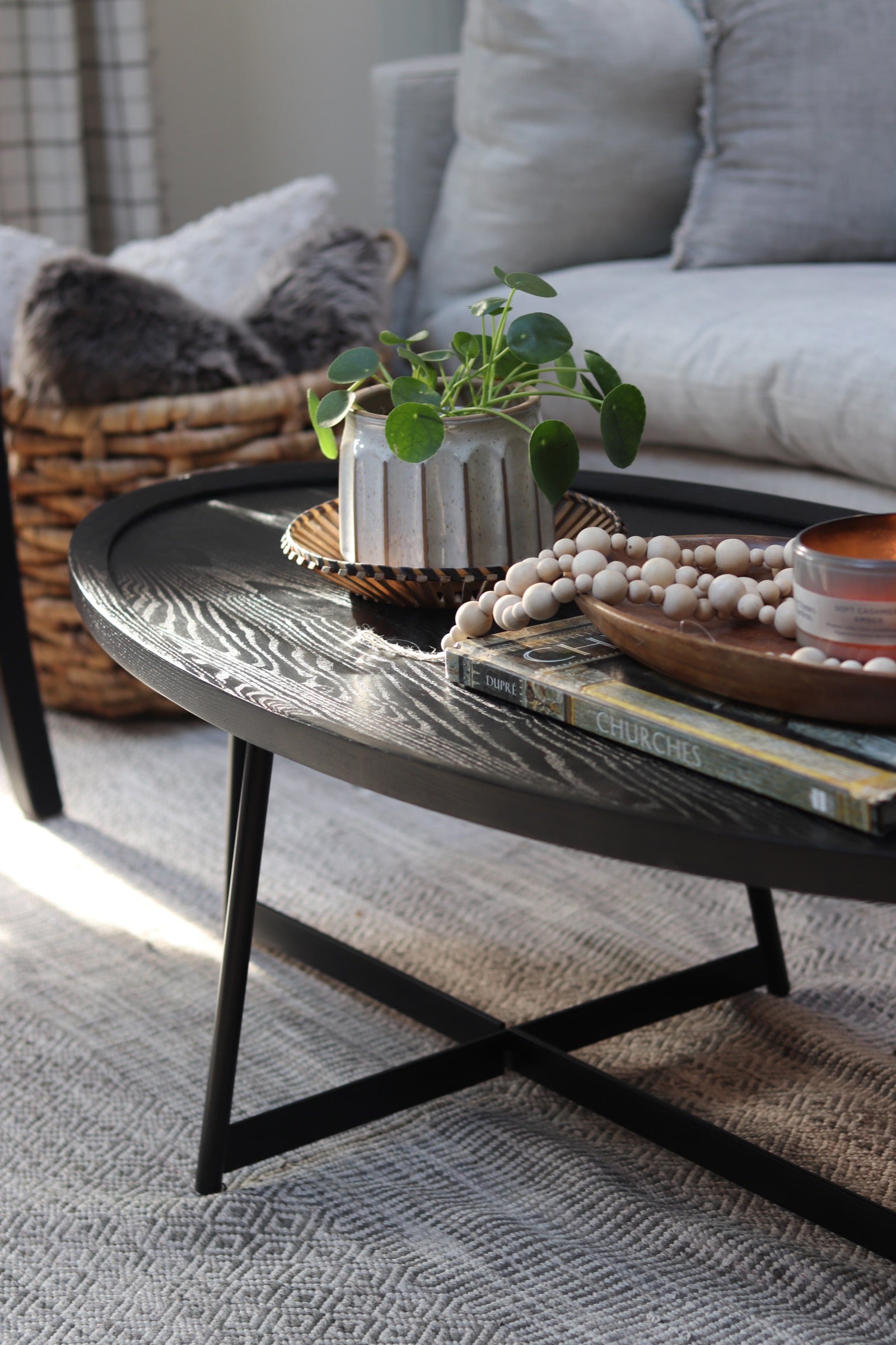 A black oval wooden coffee table holds a small potted house plant, books and decorative beads. The table sits in front of a gray sofa.