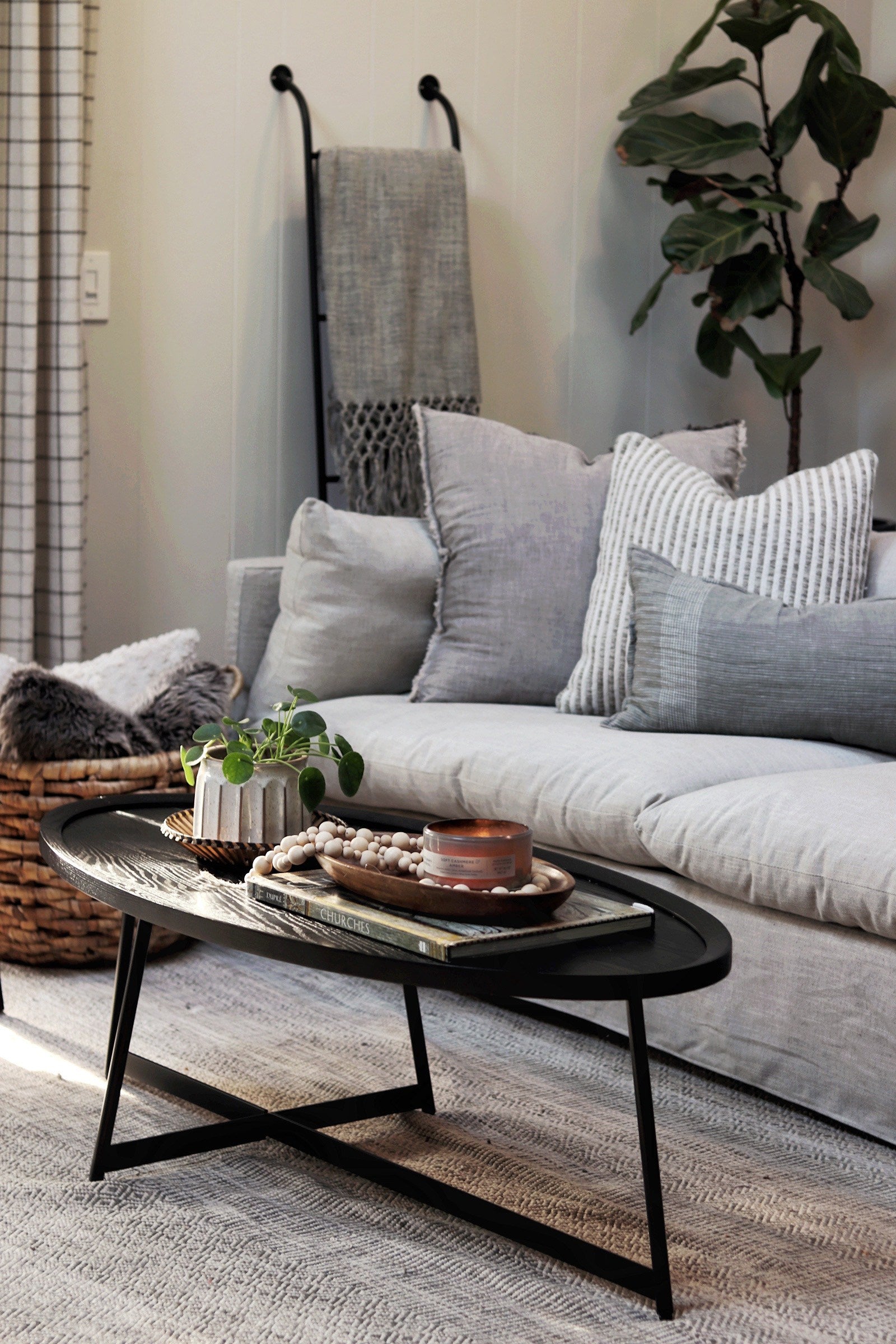 A black oval wooden coffee table holds a small potted house plant, books and decorative beads. Behind the table is a light gray slipcovered Arlen sofa with gray pillows.
