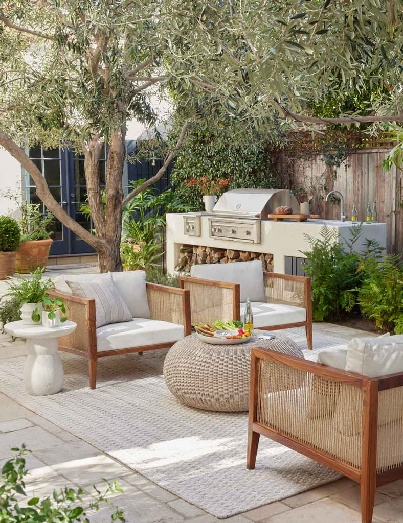 Two squared wooden accent chairs with white cushions sit on a patio with a round wicker coffee table ottoman holding snacks. Behind the seating area is an outdoor kitchen with a grill and sink.