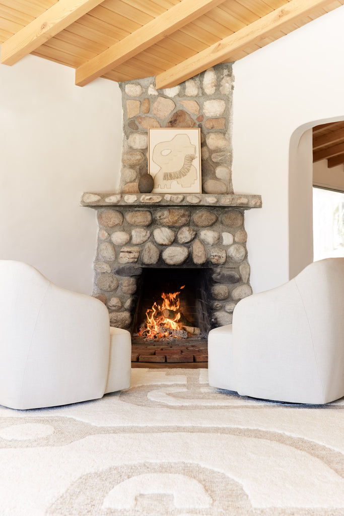 Two curved ivory accent chairs sit in front of a stacked river rock stone fireplace and atop a geometric neutral area rug.