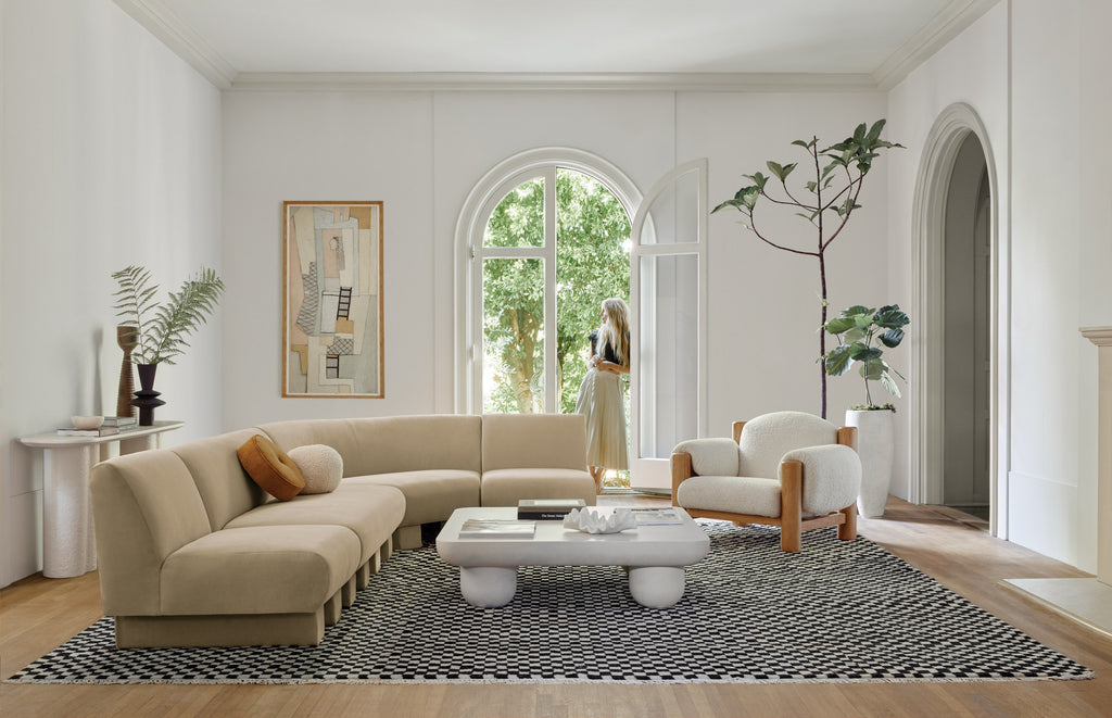 The modular Lena sectional sofa in a light taupe velvet sits across from the sherpa upholstered James accent chair and is anchored by a black and white Checkerboard rug, all designed by Sarah Sherman Samuel. The white clouded coffee table sits in the middle of the room, and the designer leans against an open door in the back.