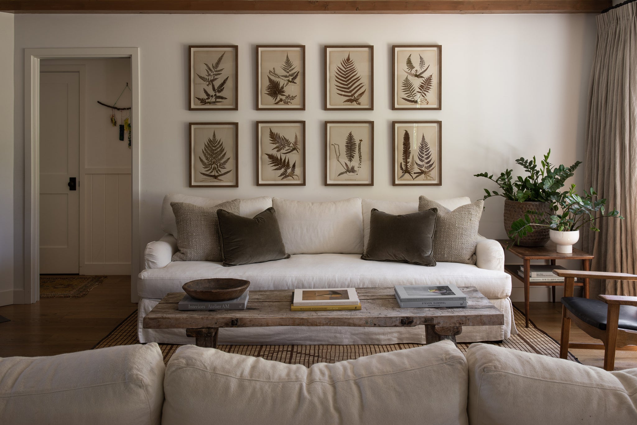 Sarah Solis' living room has two facing linen slipcovered sofas with neutral solid throw pillows. Between the couches sits a rustic wooden rectangular coffee table with books. Above a sofa are eight drawings of fern leaves and next to the sofa is a small wooden end table with two house plants.