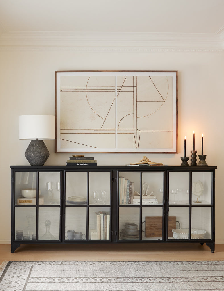 A black sideboard cabinet with clear class doors sits against a white wall. The cabinet holds decor items and a gray stone lamp sits on the left while three lit taper candles sit on the right. Above the buffet is a geometric line drawing.