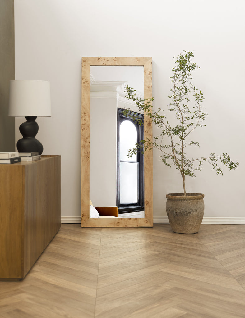 A full-length burl wood mirror leans against a wall in this entryway. Next to the mirror sits a potted small tree and a wooden sideboard cabinet.