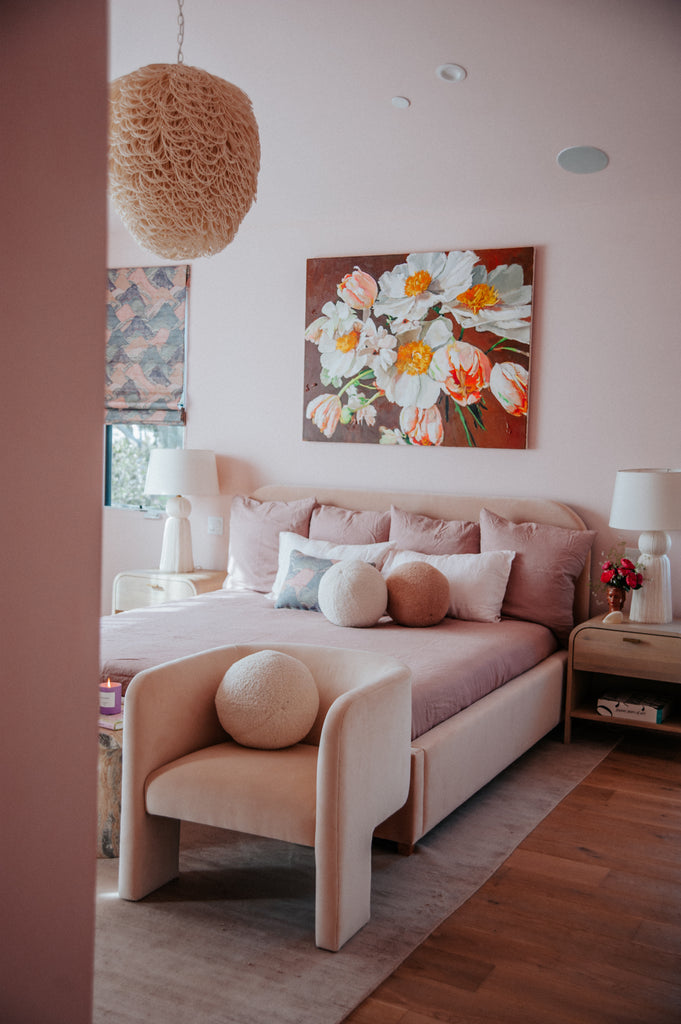 A large flower painting hangs above the pink Solene platform upholstered bed with pink bedding and sphere throw pillows. At the end of the bed is a curved pink velvet chair with a matching pink boucle sphere pillow.