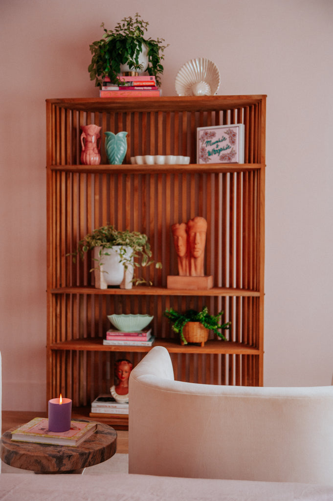 A curved wooden bookcase with slats on the back holds house plants, books, and vases.