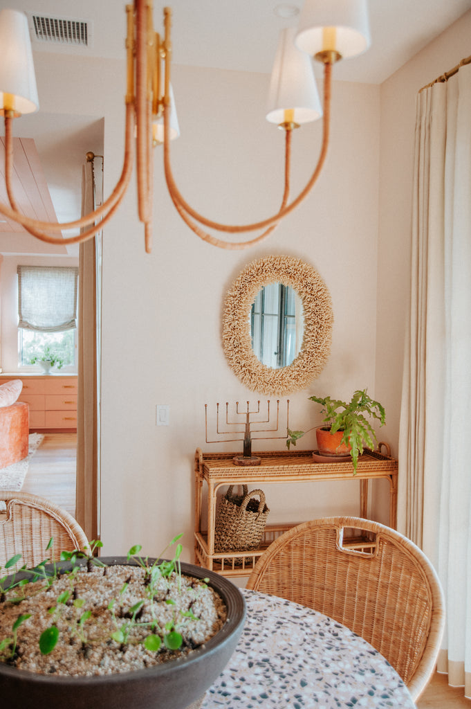 A low bowl full of growing sprouts sits in the middle of a stone kitchen table flanked by wicker arm chairs. Behind it hangs an oval framed mirror above a rattan bar cart and a modern square Menorah.