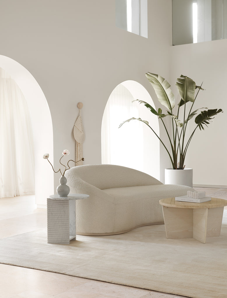 The Lyric teardrop-shaped woven wall hanging made by Candice Luter hangs behind a white boucle curved sofa flanked by a large potted plant. A light wood round coffee table and small round marble side table flank the sofa.