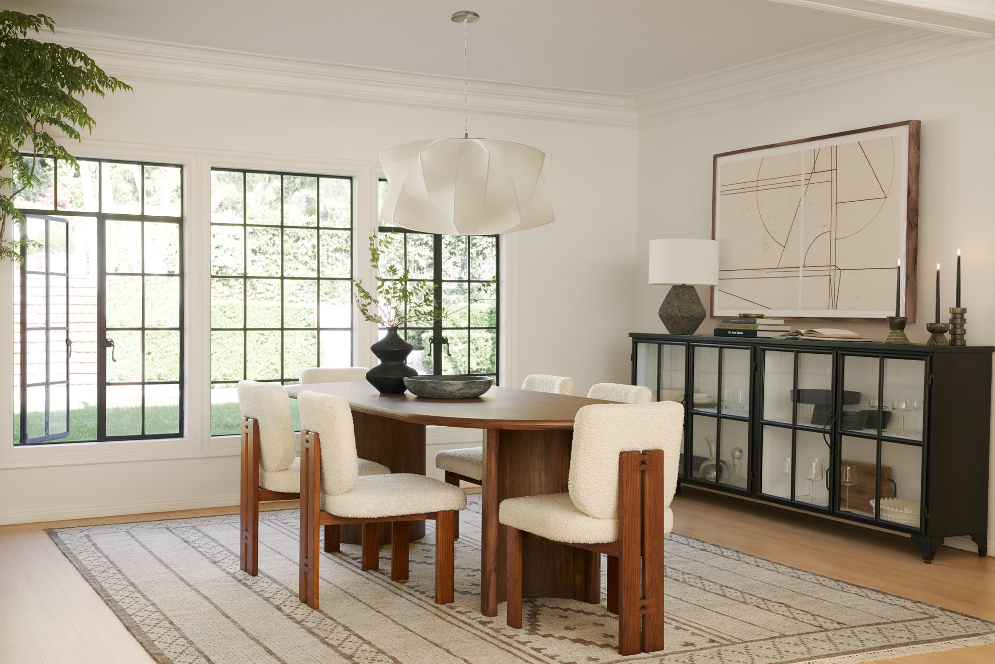 Six white boucle dining chairs with wooden backs and legs sit at an oval medium brown wood dining table. The seating sits on a white and gray geometric area rug and a modern white sculptural chandelier hangs above. A black sideboard buffet is off to the side.