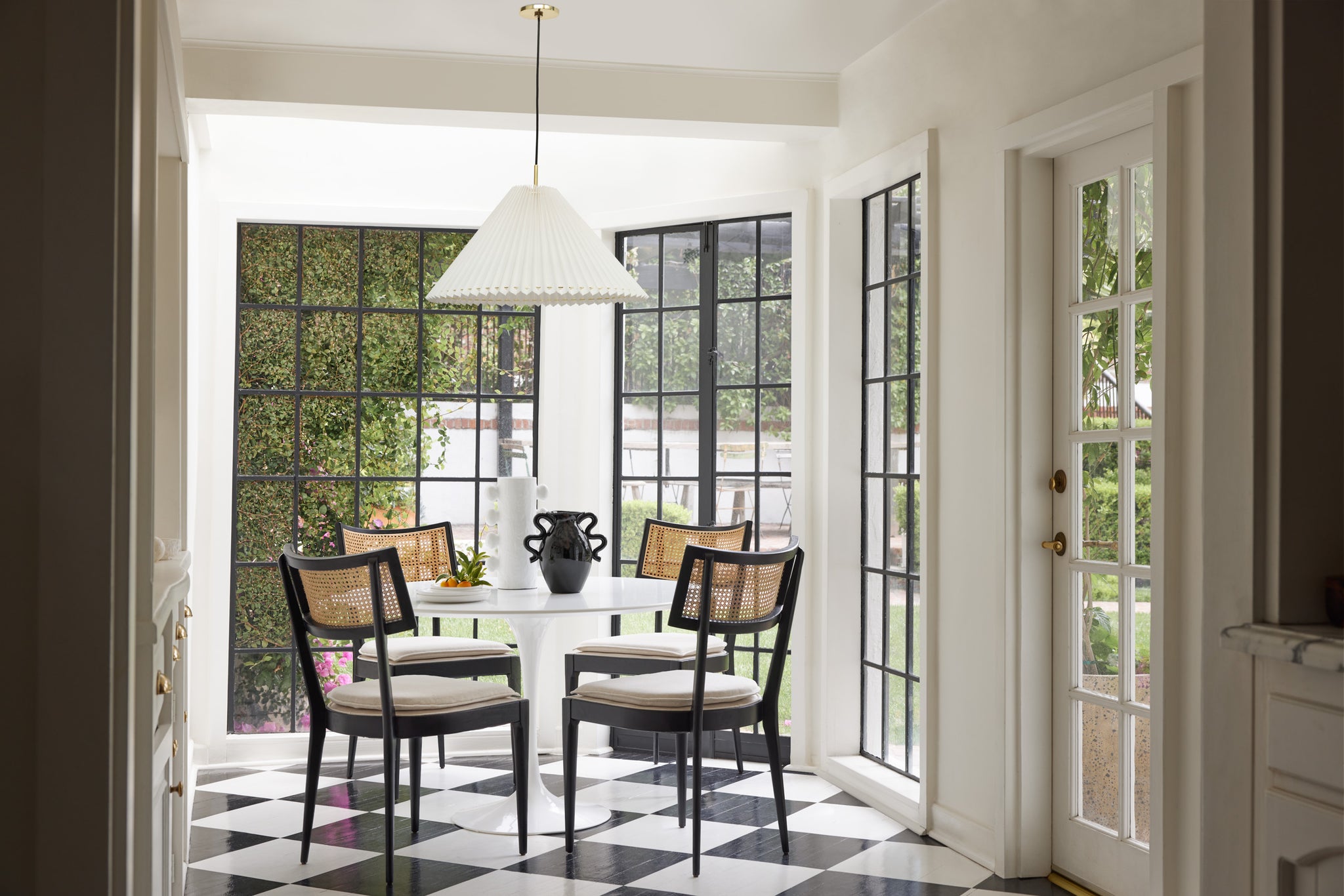 Four black dining chairs with rattan backs and upholstered seats surround a white round table in a kitchen dining nook. A white cylindrical vase and shiny black vase sit on top of the table.